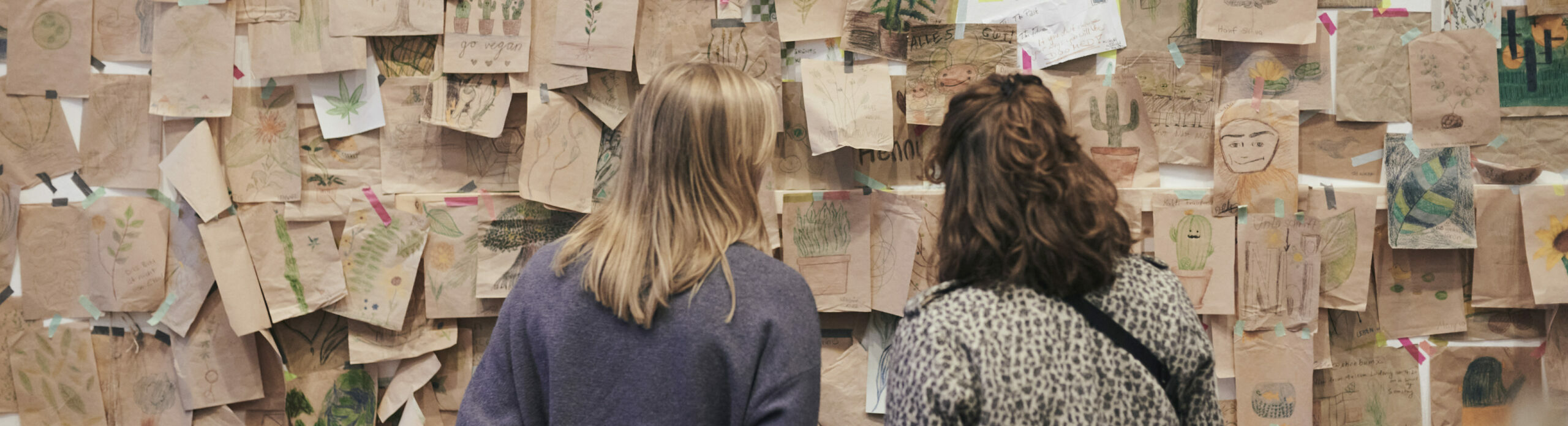 Zwei Frauen betrachten eine Wand mit selbstgemalten Bildern aus einer Ausstellung