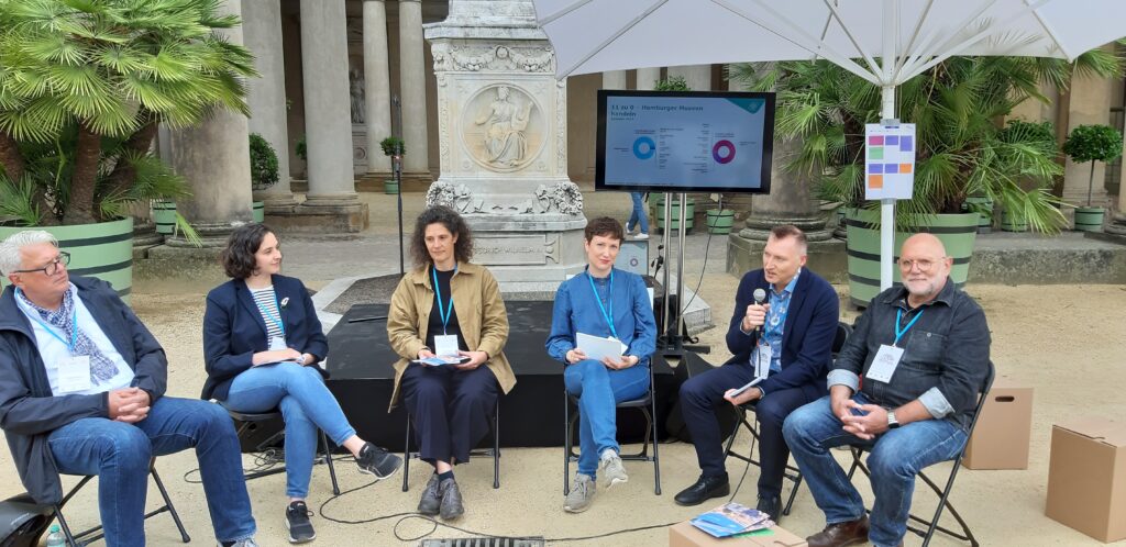 Panel-Teilnehmer:innen v.l. Thomas Hermann, Aurelia Weritz, Ulrike Fischer, Hanna Belz, David Vuillaume, Michael John
© 414films Sechs Personen sitzen auf Stühlen nebeneinander vor einer historischen Schlossfassade. Hinter ihnen ein Bildschirm mit einer Grafik und Blumenkästen mit Palmen.