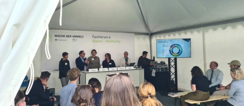 Fachforum auf der Woche der Umwelt. Fünf Personen stehen hinter einem langen Stehtisch. Daneben ist ein Bildschirm mit einer Grafik. Im Vordergrund sieht man das Publikum, das auf Bierbänken sitzt.