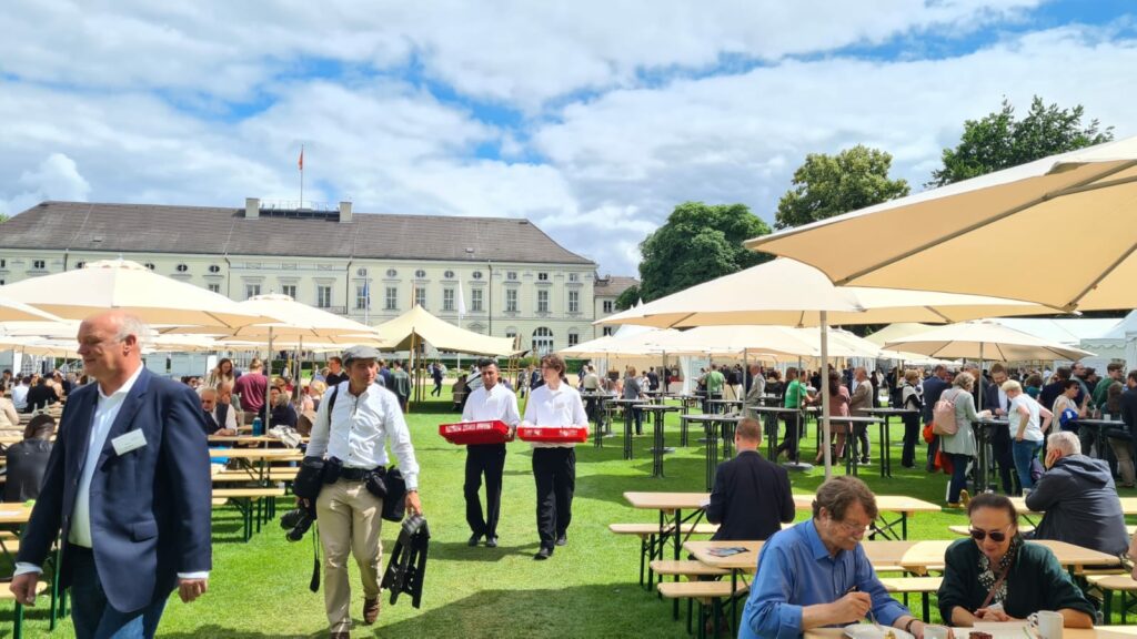 Sommerliche Veranstaltung mit Biertischen und Sonnenschirmen im Schlosspark von Bellevue. Im Hintergrund sieht man die Fassade des Schlosses.