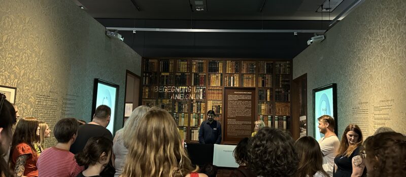 ©Rautenstrauch-Joest-Museum Köln (RJM) Eine Gruppe Menschen steht in einem Ausstellungsraum. Am anderen Ende eine Wand mit Büchern.