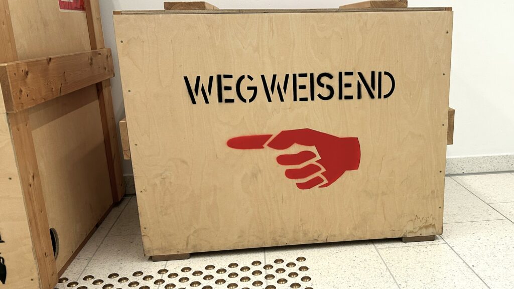 Eine Transportkiste aus Holz mit der Beschriftung "Wegweisend" und ein rotes Symbol einer Hand mit ausgestrecktem Zeigefinger. Auf dem Boden ist angeschnitten ein Blindenleitsystem zu erkennen.